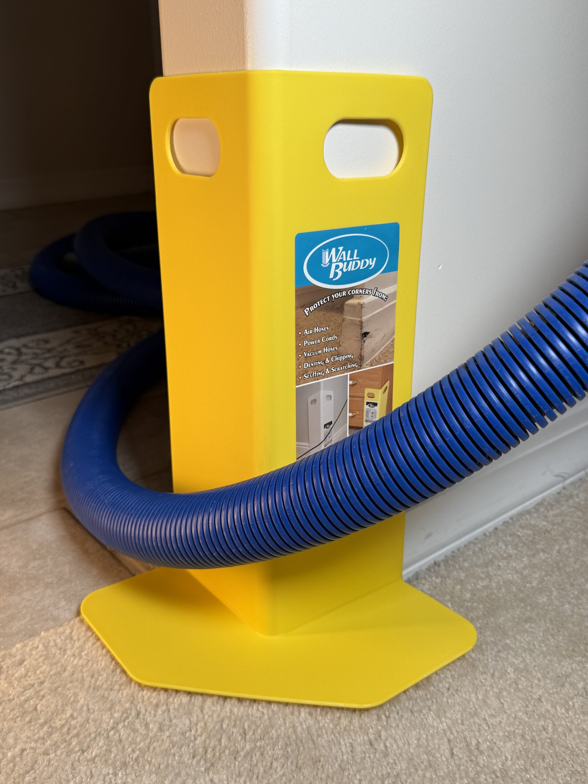 A yellow wall corner protector designed for safeguarding corners and floors. A blue vacuum hose is coiled around it, indicating its use in home maintenance.